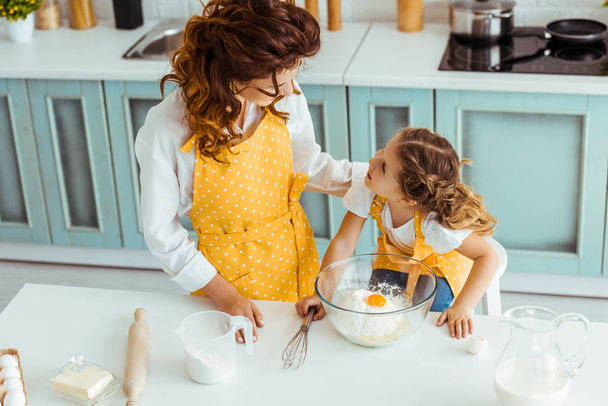 Mutter und Tochter in Schürzen schauen sich beim gemeinsamen Kochen in der Küche an - Foto, Bild