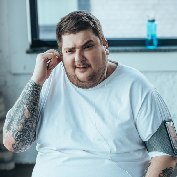 overweight man in smartphone armband looking at camera while putting on earphones at gym - Foto, Bild