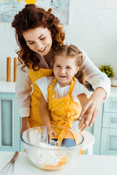 happy mother with excited daughter standing together near bowl with eggs and flour in kitchen - Foto, imagen