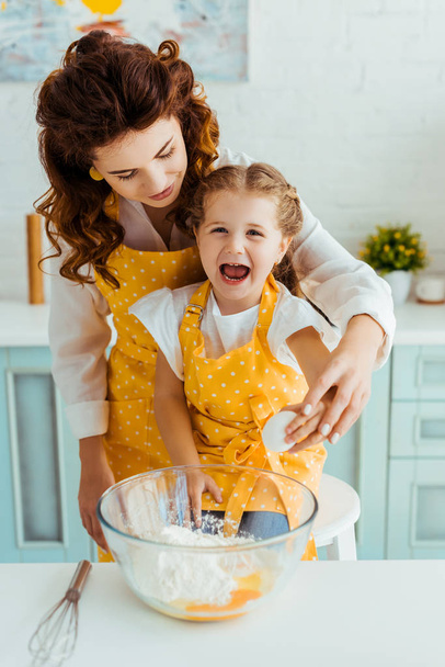 moeder en opgewonden dochter Smashing ei samen in de kom met meel in de keuken - Foto, afbeelding