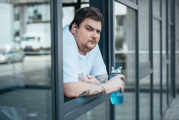 hombre obeso con auriculares y teléfono inteligente sosteniendo la botella deportiva y mirando a través de la ventana en el gimnasio
 - Foto, imagen