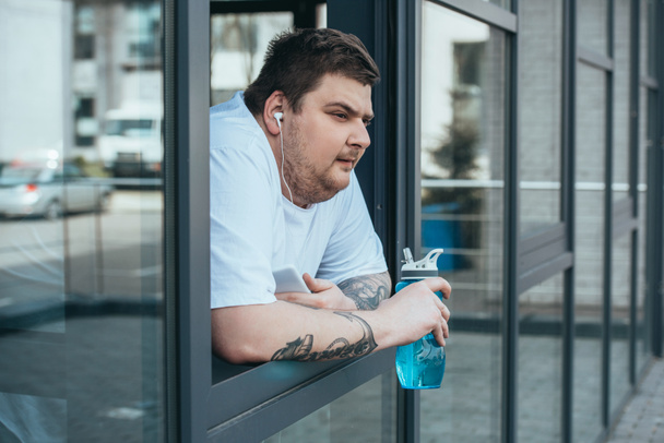 overweight man with earphones and smartphone holding sport bottle and looking out through window at gym - Photo, Image