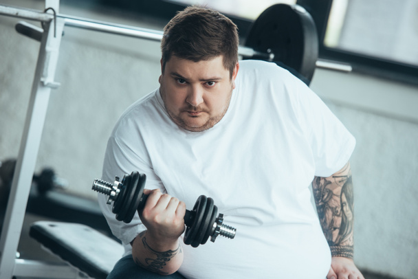 Overgewicht getatoeëerde man kijkt naar camera en training met halter in het sportcentrum - Foto, afbeelding
