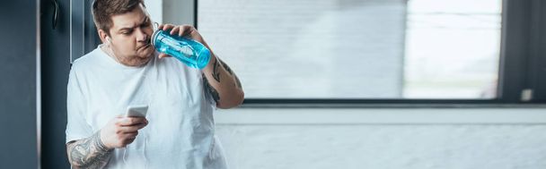 panoramic shot of overweight tattooed man using smartphone and drinking water from sport bottle at gym - Foto, Imagen