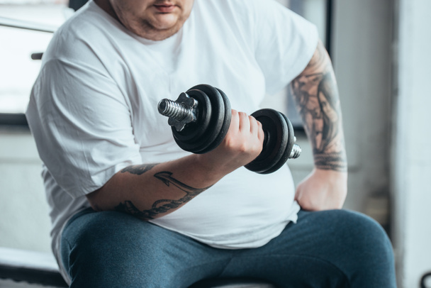 cropped view of Overweight tattooed man sitting and training with dumbbell at gym - Photo, Image