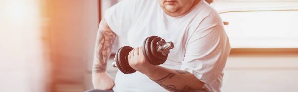 Panoramaaufnahme eines übergewichtigen tätowierten Mannes beim Training mit einer Hantel im Fitnessstudio mit Sonnenlicht - Foto, Bild
