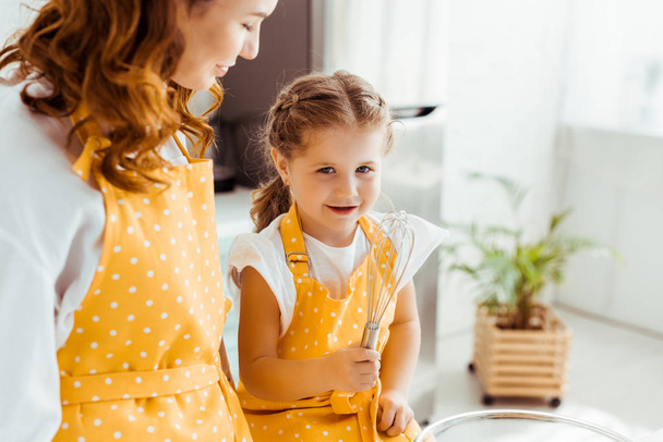 mother looking at cute daughter in polka dot apron with balloon whisk - 写真・画像