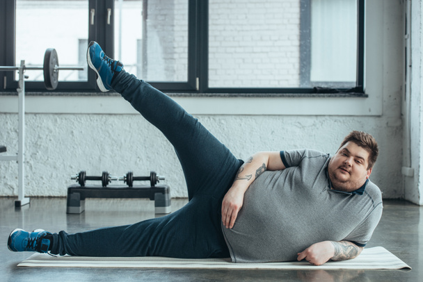 толстый татуированный мужчина лежит на коврике для фитнеса и растягивает ноги в спортзале
 - Фото, изображение