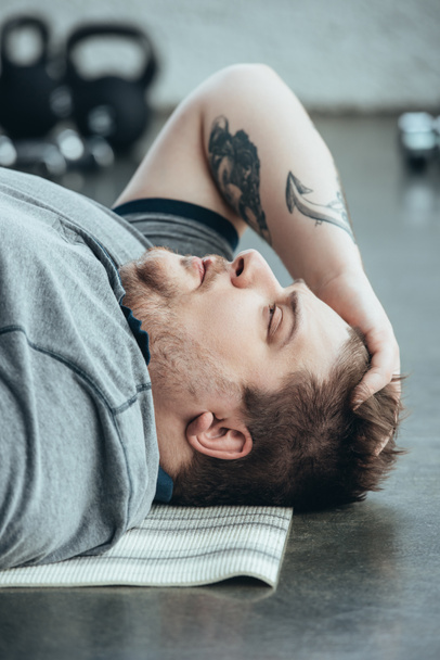 виснажений татуйований чоловік з надмірною вагою у сірій футболці, що лежить на фітнес-матусі у спортивному центрі
 - Фото, зображення