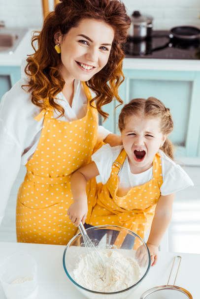overhead view of mother and excited daughter in polka dot aprons mixing flour and eggs with balloon whisk in bowl - Foto, Imagem