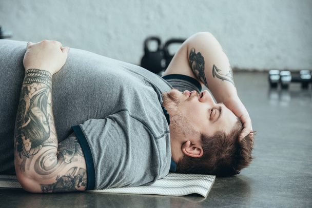 κουρασμένος υπέρβαρων τατουάζ άντρας με γκρι t-shirt που βρίσκεται στο χαλάκι Fitness στο αθλητικό κέντρο - Φωτογραφία, εικόνα