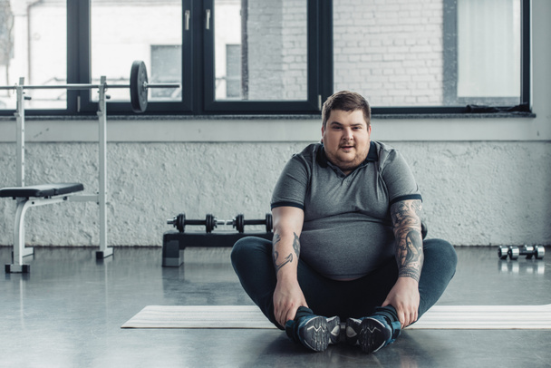 hombre tatuado con sobrepeso sentado y mirando a la cámara durante el ejercicio de estiramiento en el gimnasio
 - Foto, Imagen