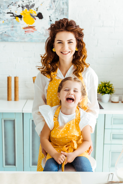 happy mother and laughing daughter in polka dot yellow aprons in kitchen - Photo, Image