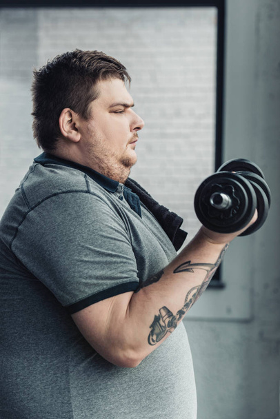 боковой вид на человека с татуировкой ожирения, тренирующегося с гантелями в спортивном центре
 - Фото, изображение