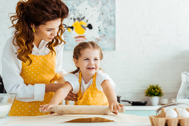 happy daughter in yellow polka dot apron rolling out dough on baking parchment paper next to mother - 写真・画像
