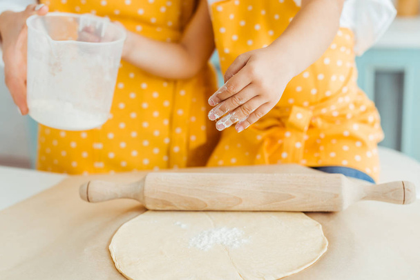 cropped view of mother and daughter in yellow polka dot aprons holding measuring jug with flour near dough and wooden rolling pin on baking parchment paper - Photo, Image