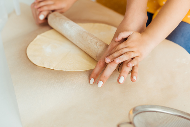 partial view of mother and daughter rolling out dough with wooden rolling pin on baking parchment paper - Valokuva, kuva