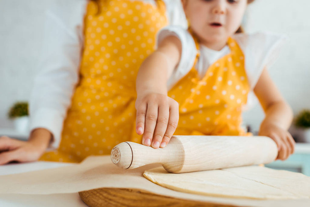 cropped view of mother and excited daughter rolling out dough with wooden rolling pin on baking parchment paper - Photo, Image
