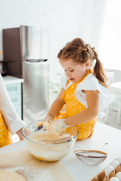 excited kid in polka dot yellow apron mixing dough in bowl with hands - Φωτογραφία, εικόνα