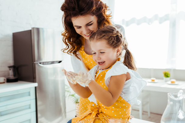 happy mother looking at laughing daughter with dirty hands in dough - Фото, изображение
