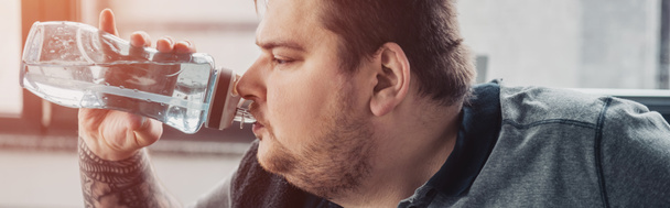 panoramic shot of overweight tattooed man drinking water from sport bottle at gym with sunlight - Fotoğraf, Görsel