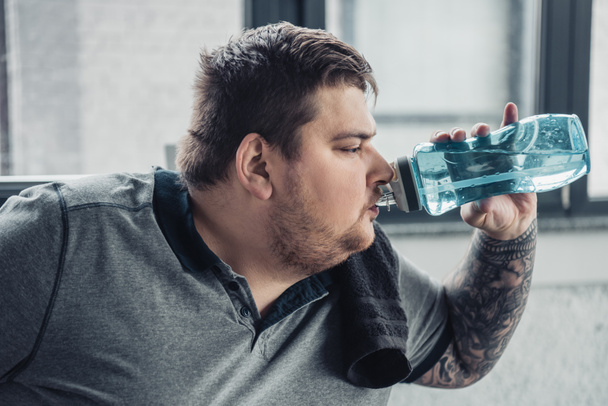 надмірна вага татуйованого чоловіка п'є воду зі спортивної пляшки в спортивному центрі
 - Фото, зображення