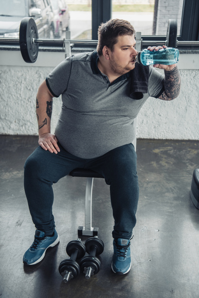 Надмірна вага татуйованого чоловіка п'є воду зі спортивної пляшки після тренування з гантелями у спортивному центрі
 - Фото, зображення