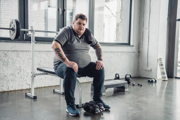 cansado Hombre obeso sentado en el banco y mirando a la cámara después de hacer ejercicio con pesas en el gimnasio
 - Foto, imagen