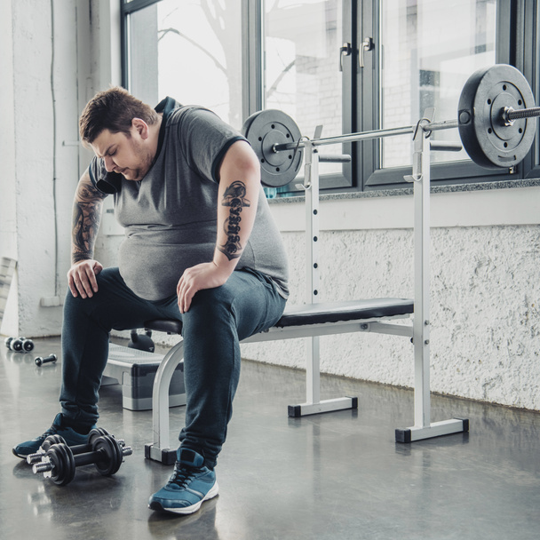cansado Hombre obeso sentado en el banco después de hacer ejercicio con pesas en el gimnasio
 - Foto, Imagen