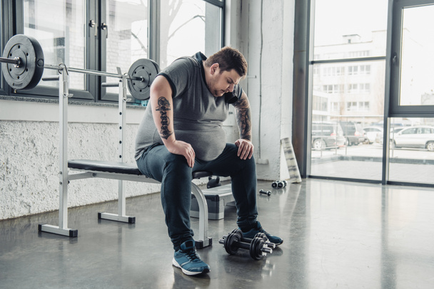 unavené nadváha muž sedící na lavičce po cvičení s činkami v tělocvičně - Fotografie, Obrázek