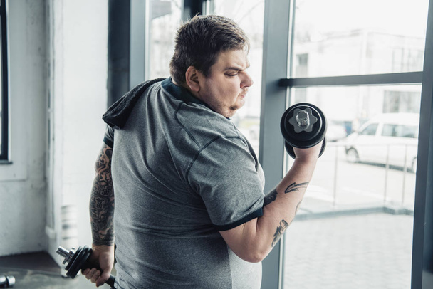 Übergewichtiger tätowierter Mann mit Handtuch trainiert mit Kurzhanteln im Sportzentrum - Foto, Bild