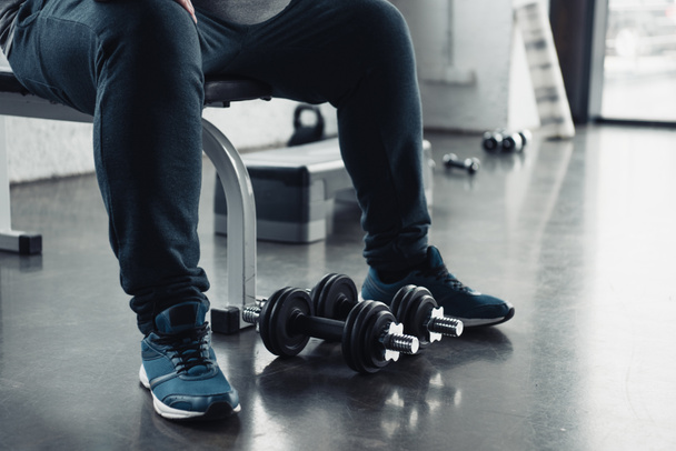cropped view of man in trainers sitting with dumbbells at sports center - Fotografie, Obrázek