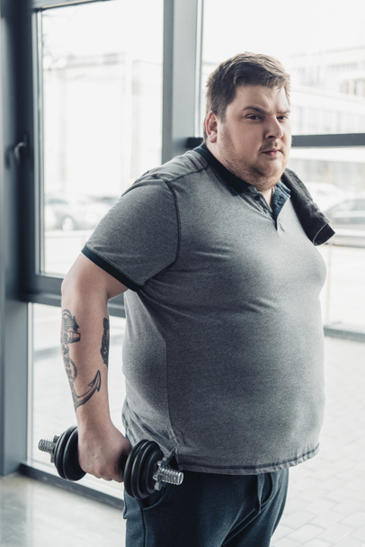 Hombre tatuado con sobrepeso en camiseta gris con toalla haciendo ejercicio con mancuerna en el centro deportivo
 - Foto, Imagen
