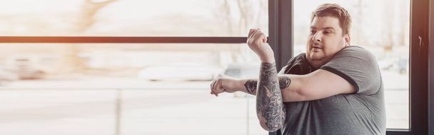 panoramic shot of overweight tattooed man stretching arms at gym with copy space - Photo, Image