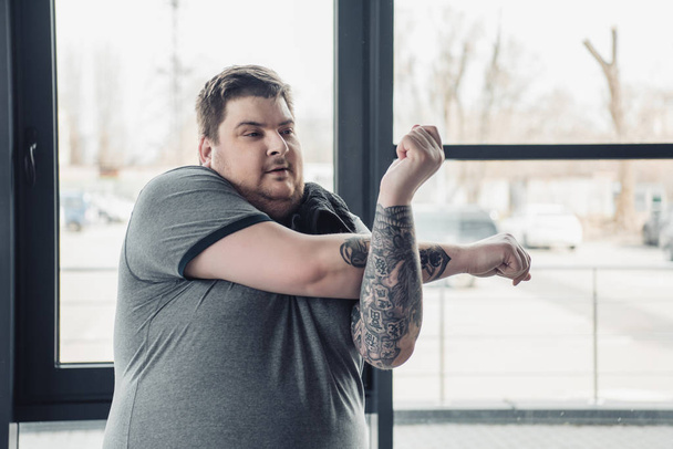 sobrepeso tatuado homem em cinza t-shirt alongamento braços no ginásio
 - Foto, Imagem