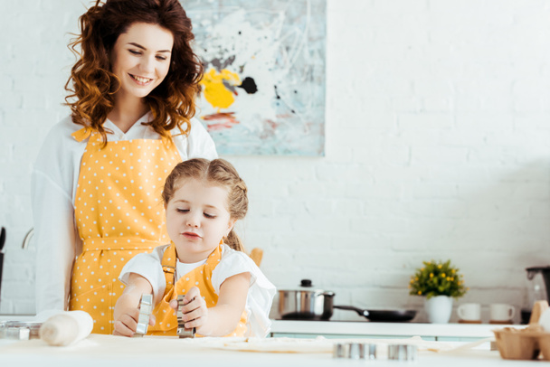 happy mother in yellow polka dot apron looking at daughter holding dough molds in kitchen - Foto, Imagem