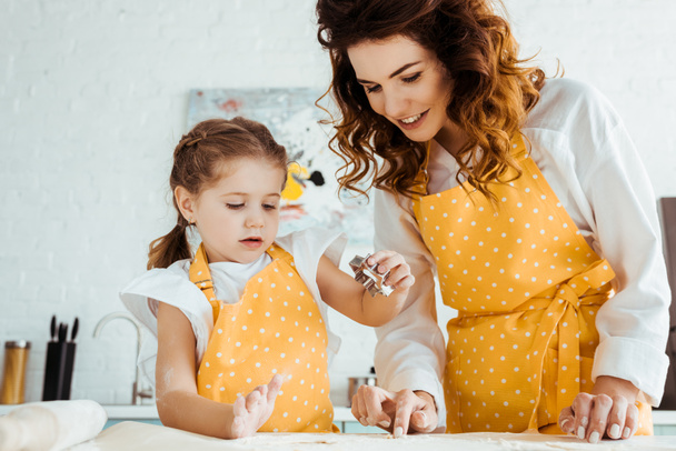 happy mother in yellow polka dot apron helping daughter using dough mold in kitchen - Valokuva, kuva