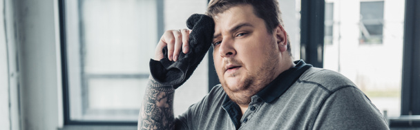 panoramische foto van moe overgewicht man kijken naar camera en vegen gezicht met handdoek in het sportcentrum - Foto, afbeelding
