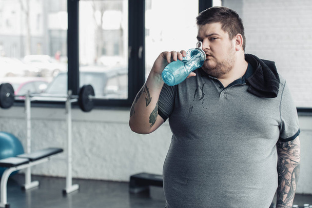 overweight tattooed man with towel drinking water from sport bottle at gym - Photo, Image