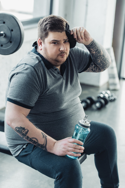 overgewicht getatoeëerd man Holding sport fles, kijken naar camera en vegen gezicht met handdoek op sportschool - Foto, afbeelding