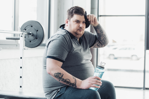 hombre tatuado con sobrepeso sosteniendo una botella deportiva y limpiando la cara con una toalla en el gimnasio
 - Foto, Imagen
