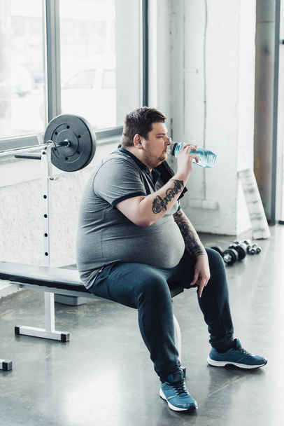 sobrepeso hombre tatuado sentado en el banco y beber agua de la botella de deporte en el gimnasio
 - Foto, Imagen