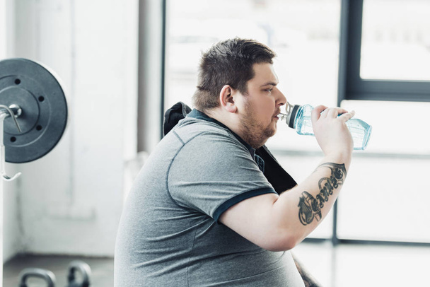 vista laterale dell'uomo tatuato sovrappeso che beve acqua dalla bottiglia sportiva in palestra
 - Foto, immagini