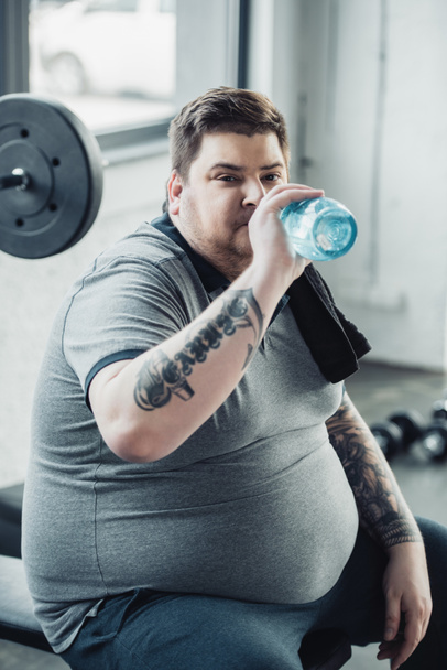 tired overweight tattooed man drinking water from sport bottle at gym - Photo, Image