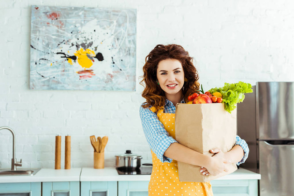 smiling woman in polka dot apron holding paper bag with fresh fruits and vegetables - Fotografie, Obrázek