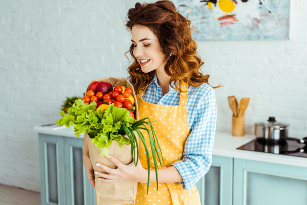 Mujer sonriente en delantal de lunares sosteniendo bolsa de papel con frutas y verduras maduras
 - Foto, Imagen