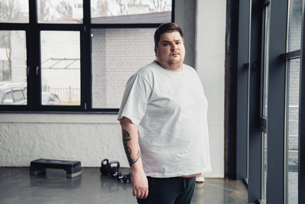 hombre tatuado con sobrepeso en camiseta blanca mirando a la cámara en el centro deportivo con espacio para copiar
 - Foto, Imagen