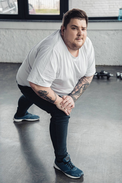 hombre tatuado con sobrepeso mirando a la cámara mientras estira las piernas en el centro deportivo
 - Foto, imagen