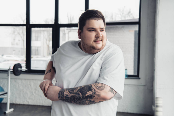 ylipainoinen tatuoitu mies valkoisessa t-paidassa venyttelemässä käsiä kuntosalilla
 - Valokuva, kuva