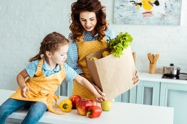 mother in polka dot apron holding paper bag while daughter sitting on table and touching apples and bell peppers - Photo, Image